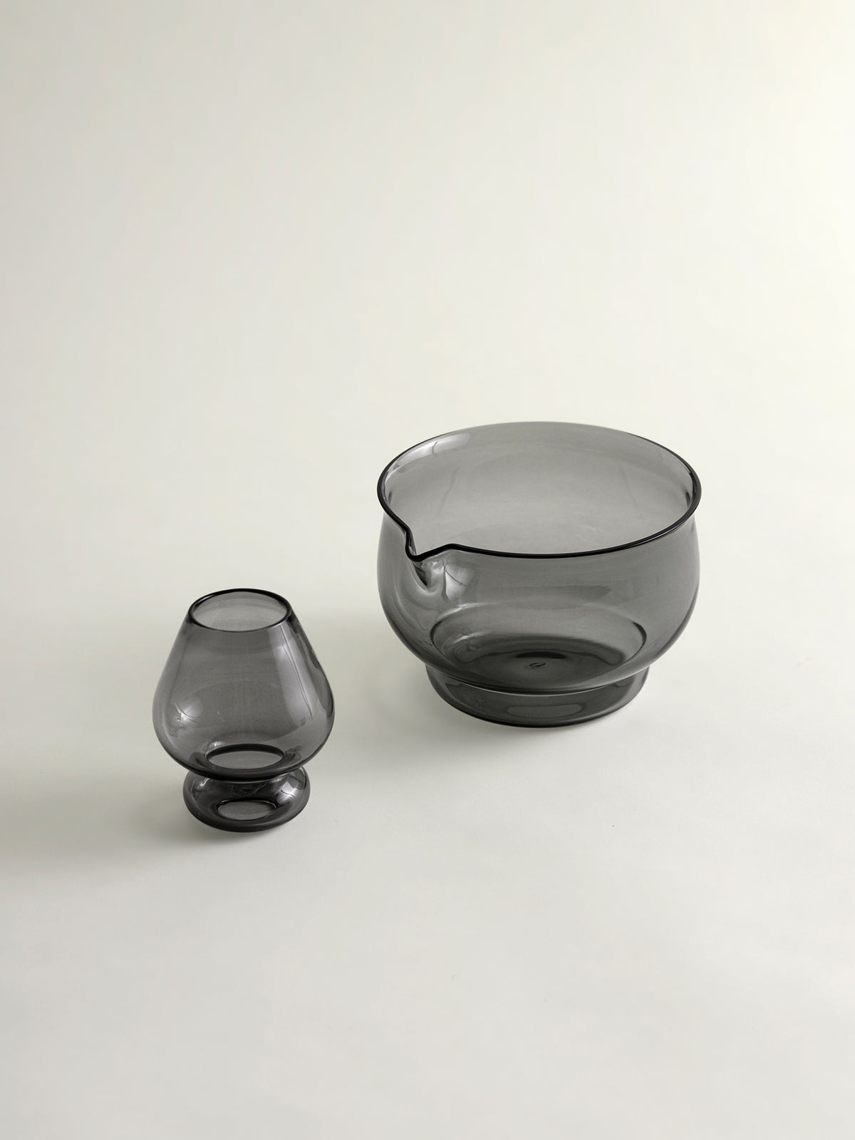 Two translucent smoky gray glass vessels on a clean off-white background, including a small whisk holder and a larger whisking bowl with a small pouring spout.