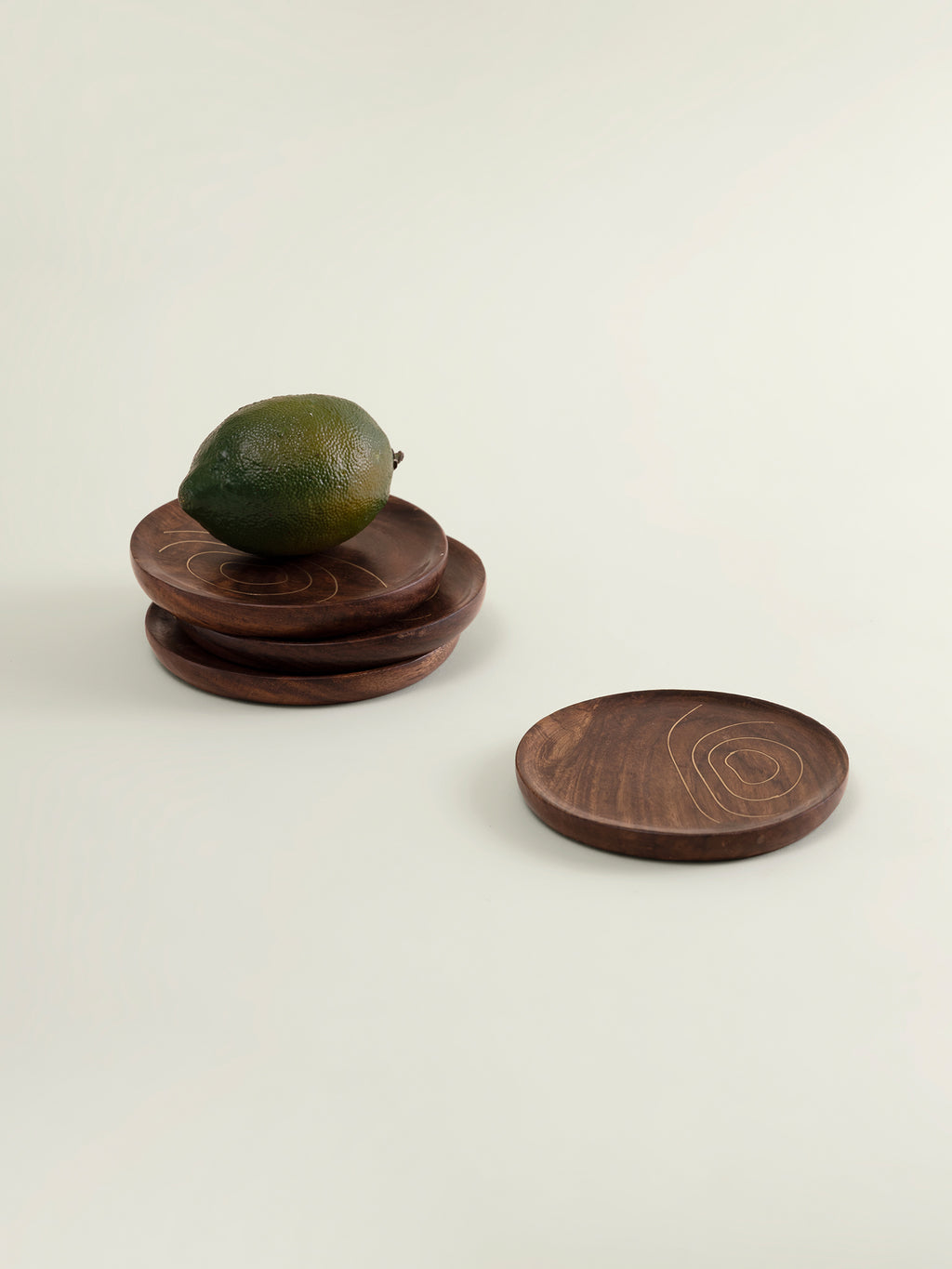 Three round dark brown wooden coasters with brass thread patterns on surface, displayed on a light cream background. One coaster is stacked on the others, with a green lime resting on top, while a second coaster sits beside them.