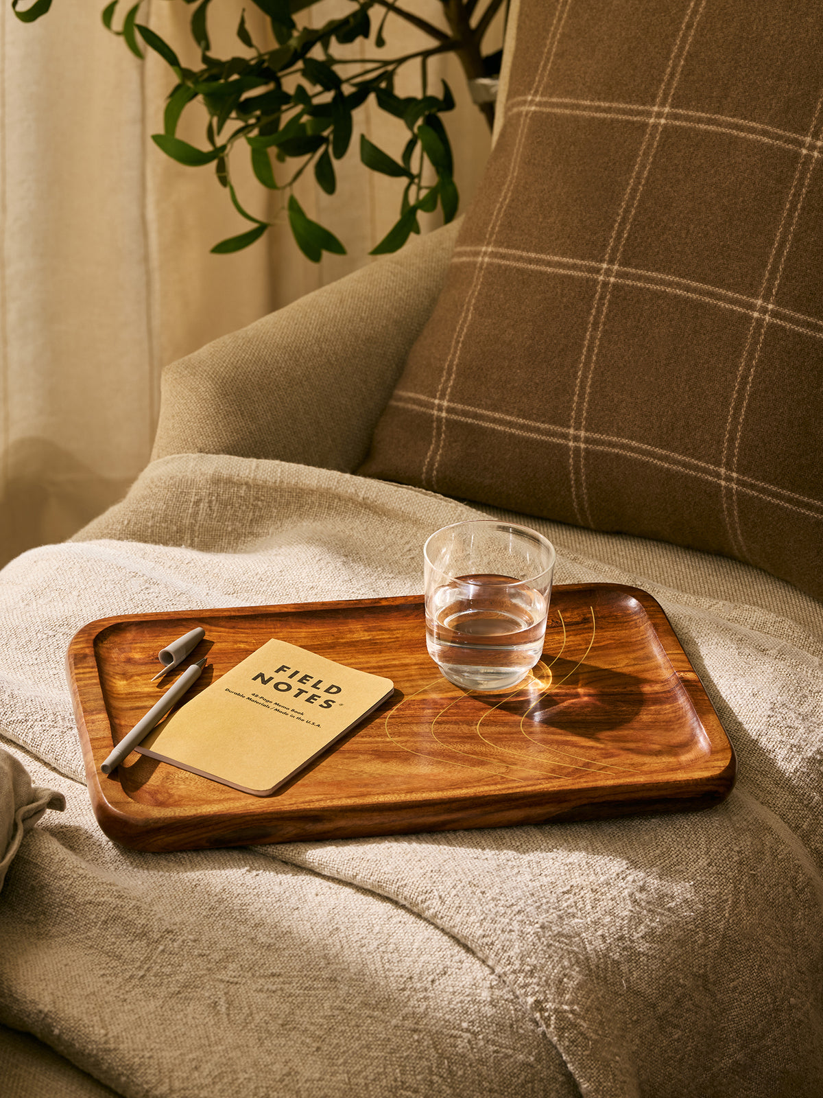 A rectangular wooden serving tray with rounded corners sits on a beige throw blanket draped over an armchair. On the tray is a small beige notebook reading 'FIELD NOTES', a pen, and a clear glass of water.