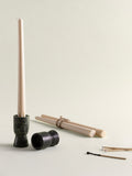 A minimalist candle holder set on a light beige background, featuring a tall cream-colored taper candle in a speckled green marble holder, a small matching holder, bundled wooden wicks, and a few matches.