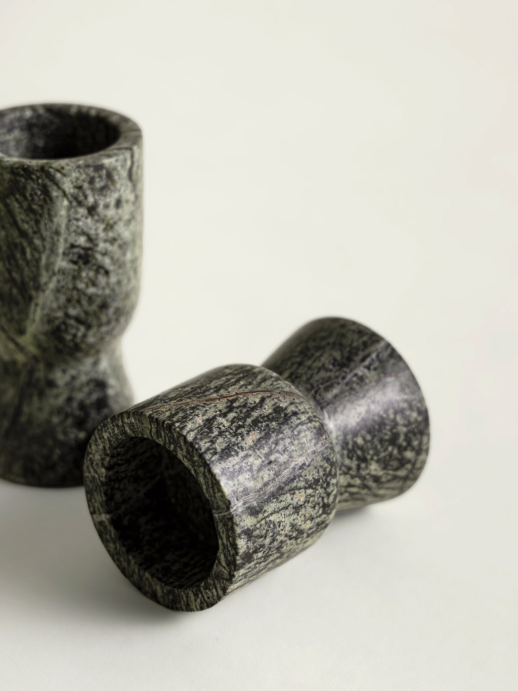 Two dark green speckled marble candle holders with a textured organic pattern, one standing upright and one lying on its side, on a light off-white background.