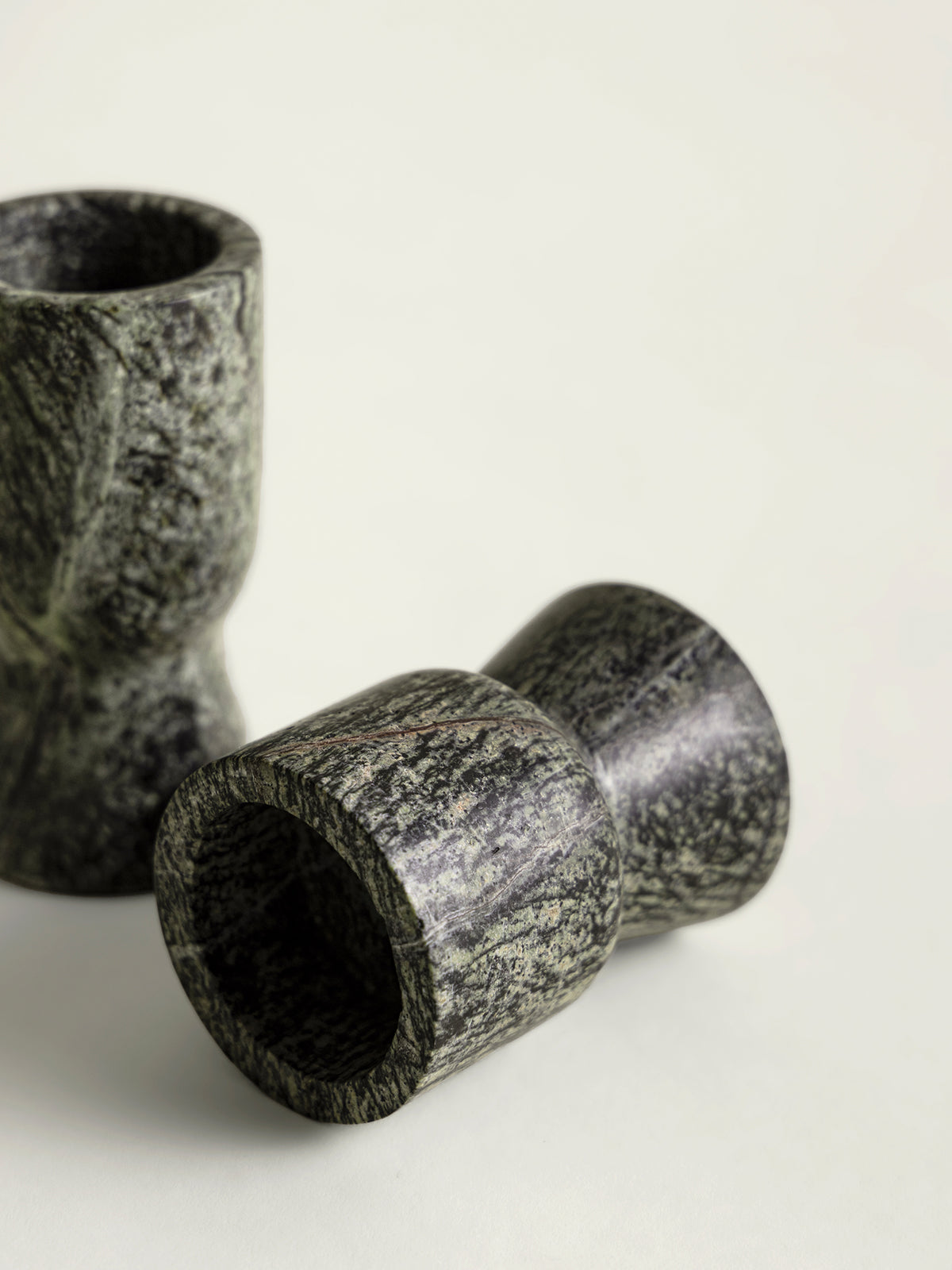 Two dark green speckled marble candle holders with a textured organic pattern, one standing upright and one lying on its side, on a light off-white background.