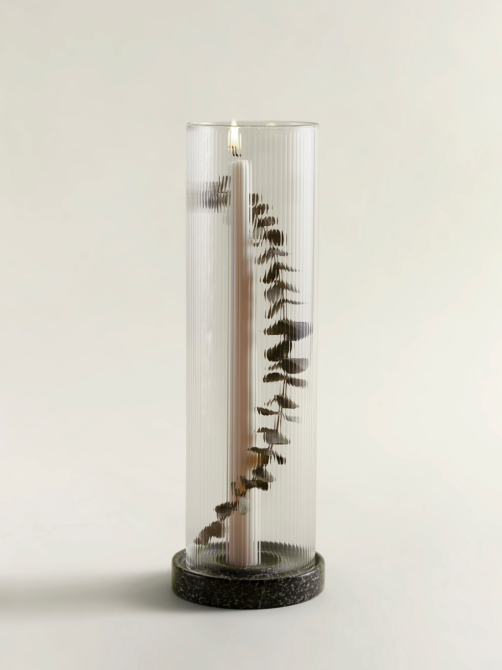 A tall ribbed clear candle lantern with a lit taper candle inside and dried eucalyptus stems arranged vertically, set on a rainforest green marble base against a plain light background.