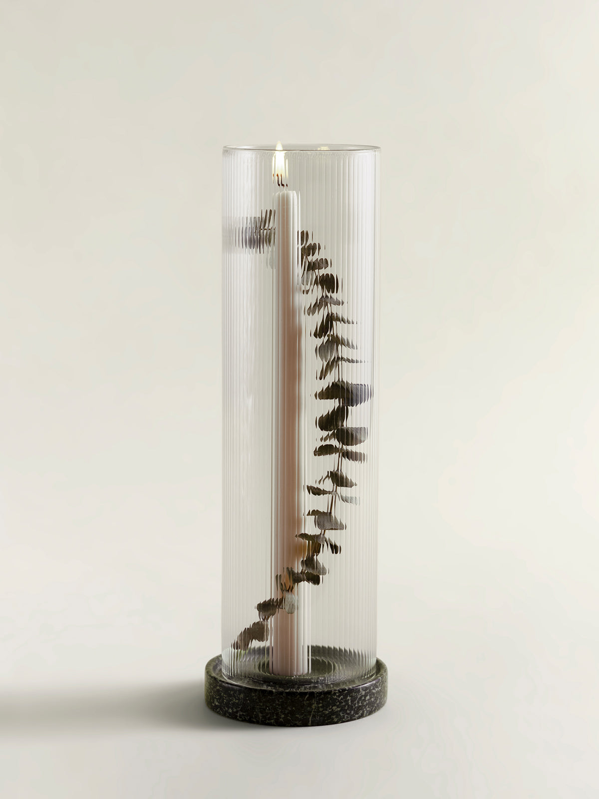 A tall ribbed clear candle lantern with a lit taper candle inside and dried eucalyptus stems arranged vertically, set on a rainforest green marble base against a plain light background.