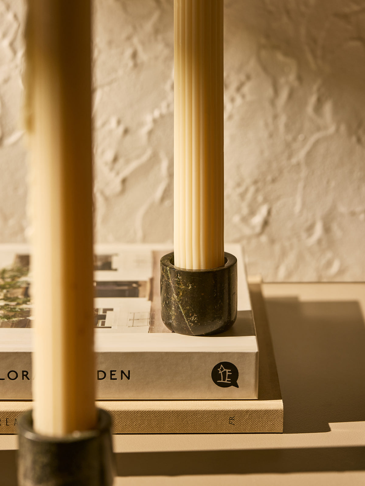 A close-up of a cream-colored ribbed candle held in a rainforest green marble holder, resting on a stack of books against a textured beige wall.