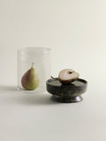 A clear ribbed glass lantern containing a pear, placed beside a green marble base holding a halved pear, on a warm off-white background.
