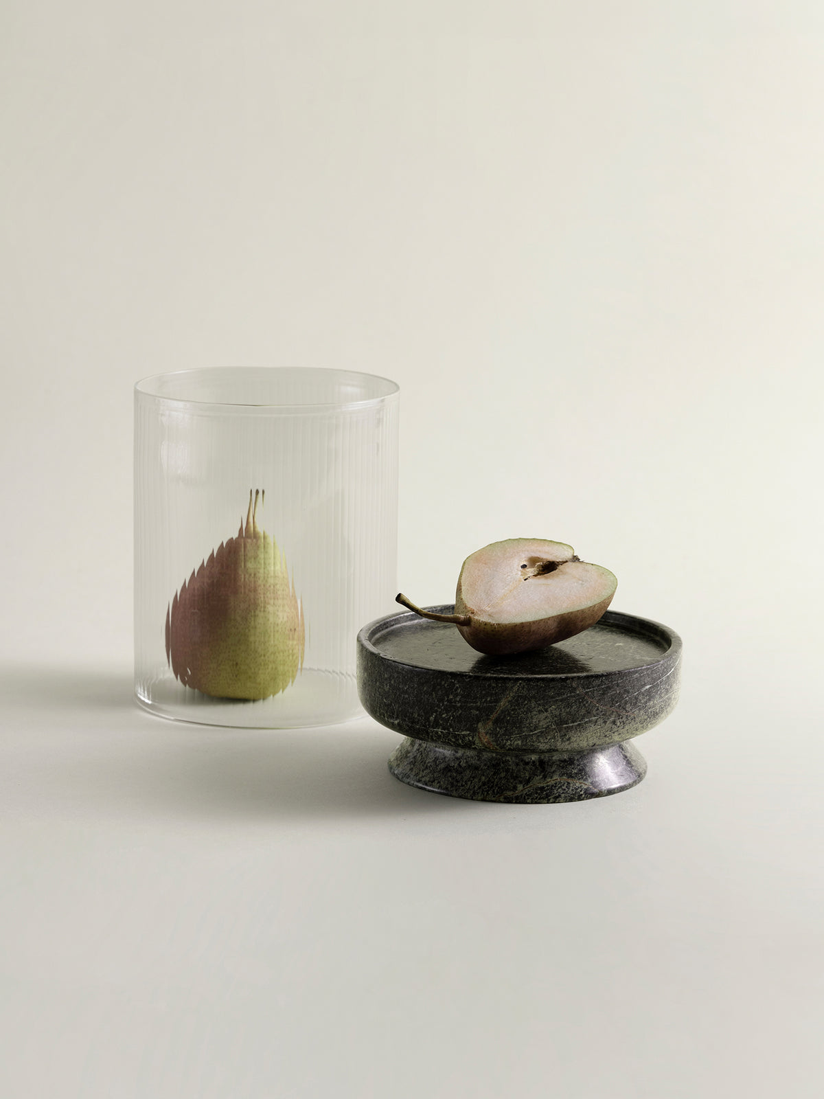 A clear ribbed glass lantern containing a pear, placed beside a green marble base holding a halved pear, on a warm off-white background.