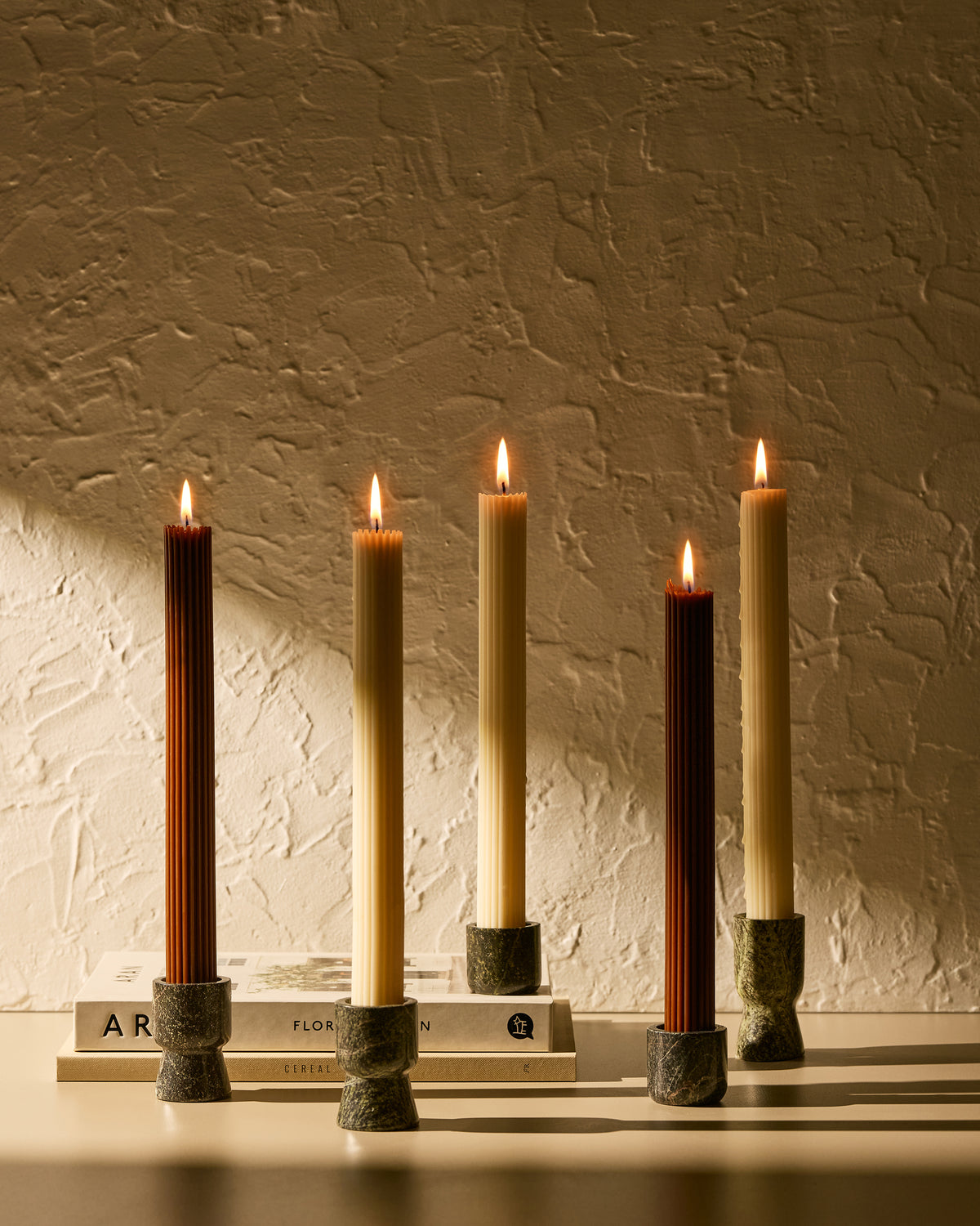 Five taper candles in rainforest green marble holders arranged on stacked books against a textured beige wall, with warm sunlight casting shadows.