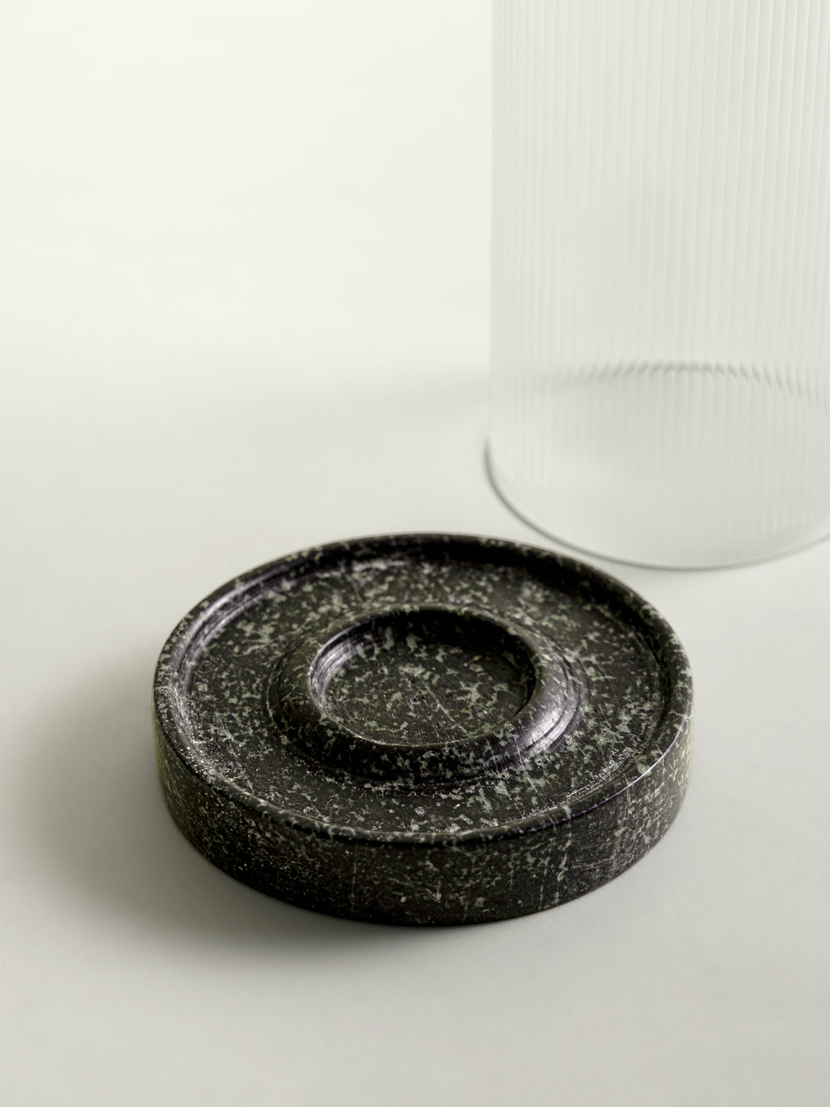 A round green speckled marble lantern base in the foreground, with a blurred clear ribbed glass lantern shade in the background on a light neutral surface.