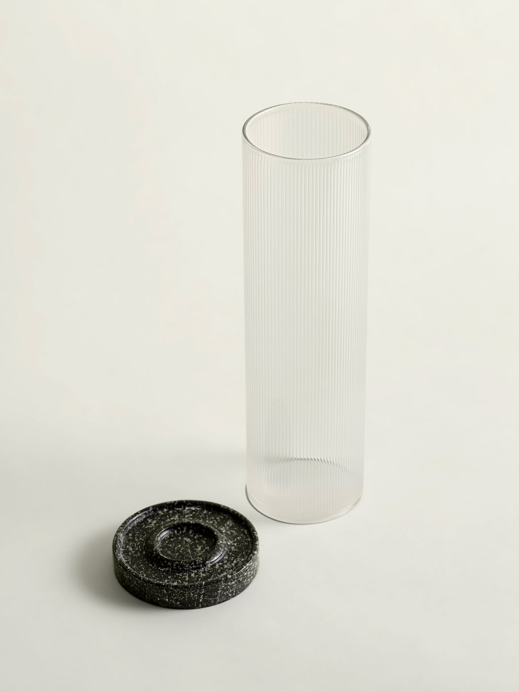 A tall, clear ribbed glass cylinder lantern top standing next to a small round green textured marble base on a clean off-white background.