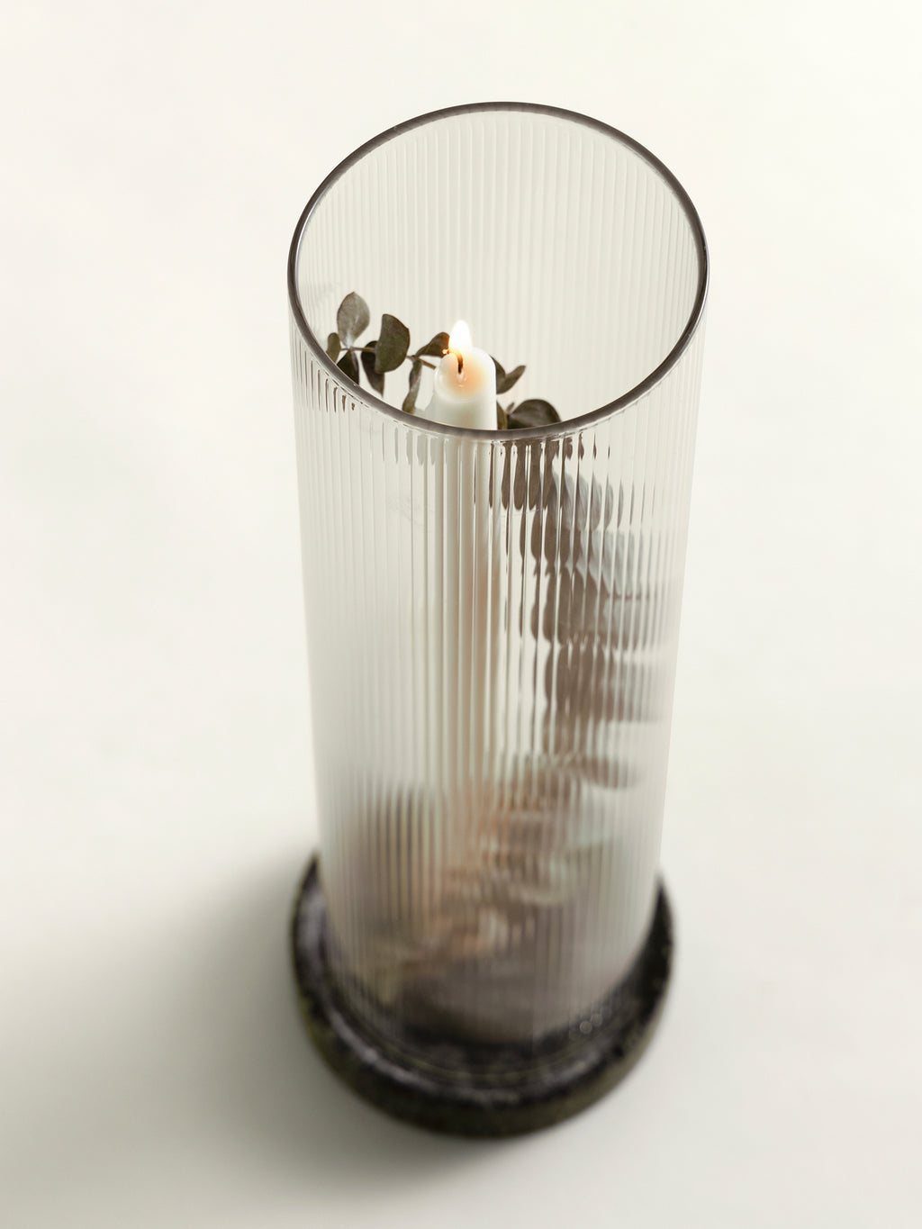 A tall ribbed clear glass lantern with a green marble base. Inside the lantern, a small white candle is surrounded by dried green leaves and stems, set against a plain white background.