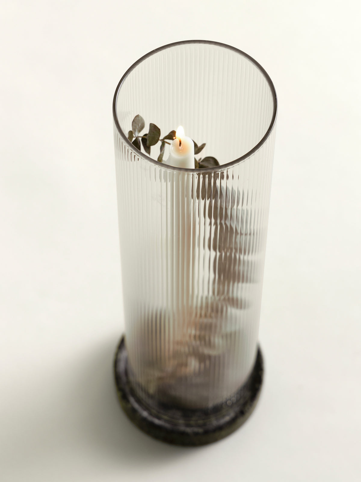 A tall ribbed clear glass lantern with a green marble base. Inside the lantern, a small white candle is surrounded by dried green leaves and stems, set against a plain white background.